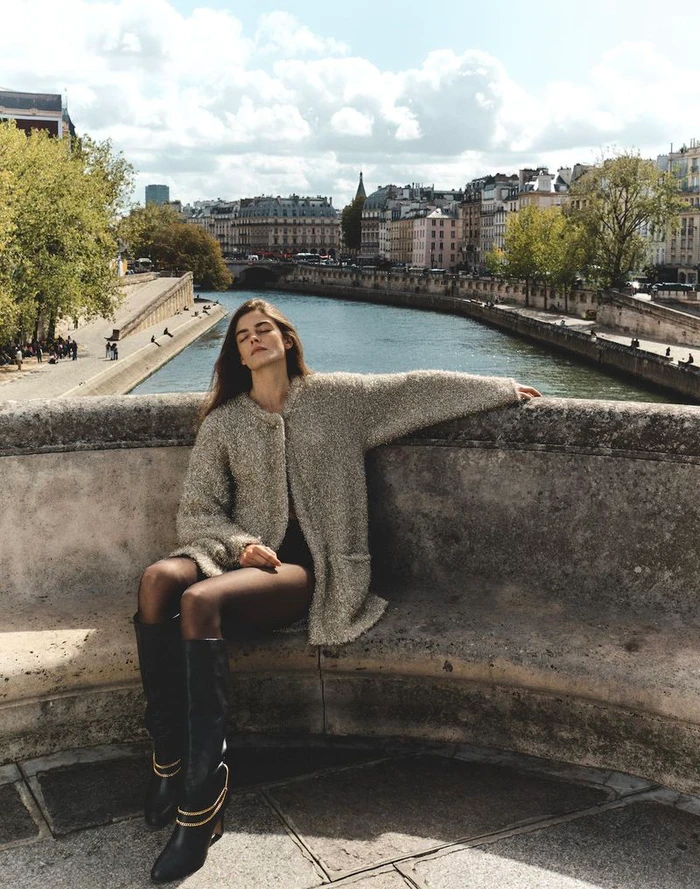 Model Merlijne Schorren seated on a stone bridge overlooking a river and city skyline for Veronica Beard