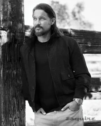 Travis Fimmel leaning against a rustic wooden fence wearing a dark jacket, black t-shirt, and watch for Esquire.