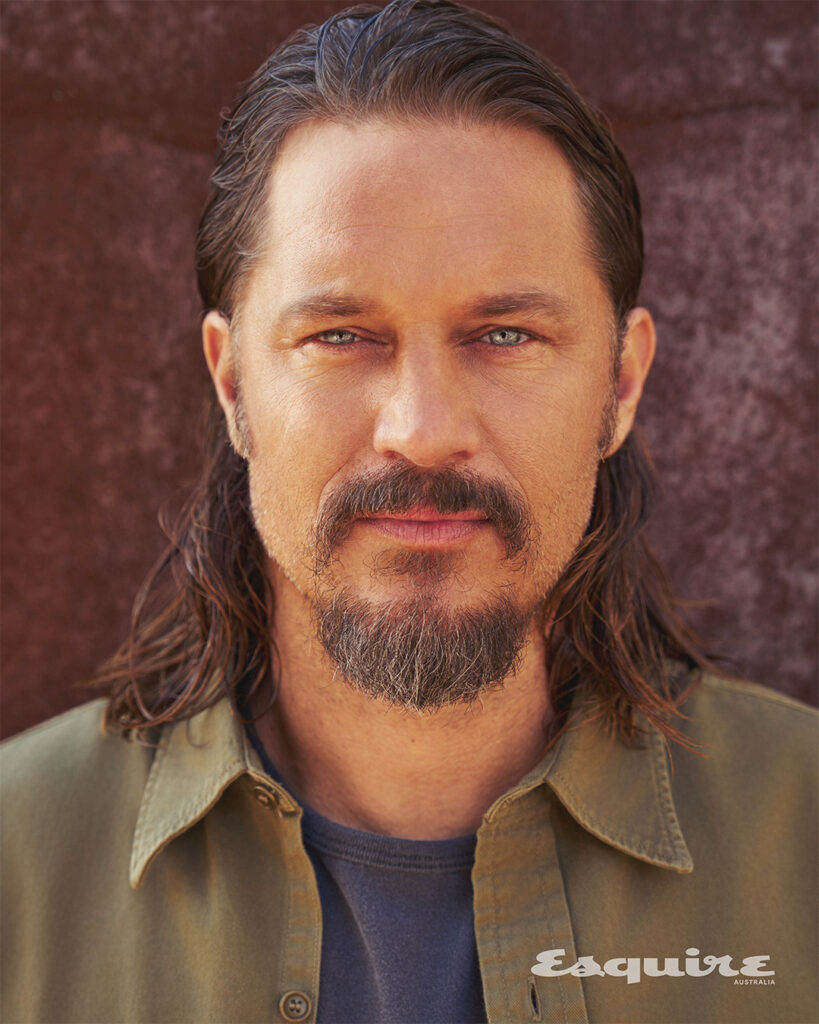 Close-up studio shot of Josh Holloway with medium-length brown hair, a mustache, and a goatee against a blurred brown background.
