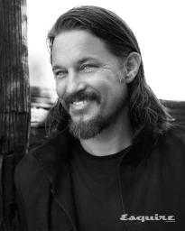 Portrait of Travis Fimmel smiling with long hair and facial hair, wearing a dark jacket outdoors for Esquire.