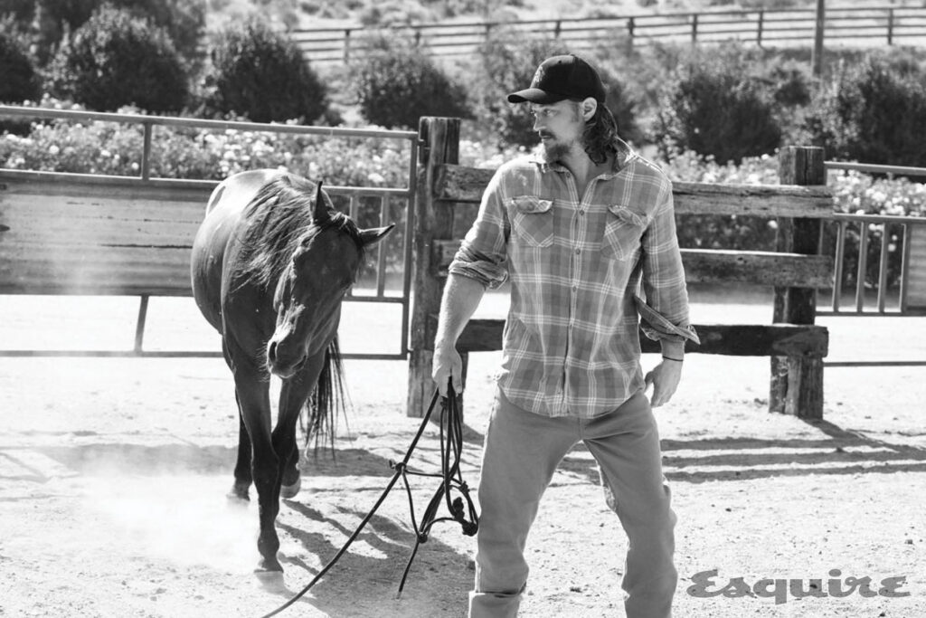Black and white shot of a man in a flannel shirt and a baseball cap leading a horse in a sandy paddock, Esquire logo in the corner.