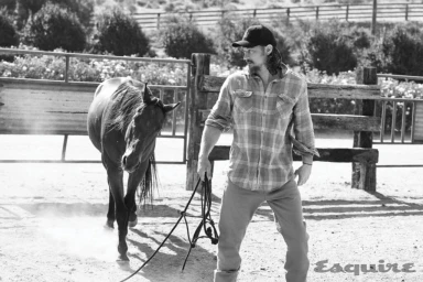 Travis Fimmel wearing a plaid shirt and cap leading a horse in a dusty corral for Esquire magazine