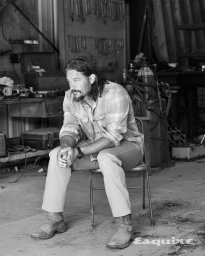 Travis Fimmel seated on a metal chair wearing a plaid shirt and cowboy boots inside a workshop garage.