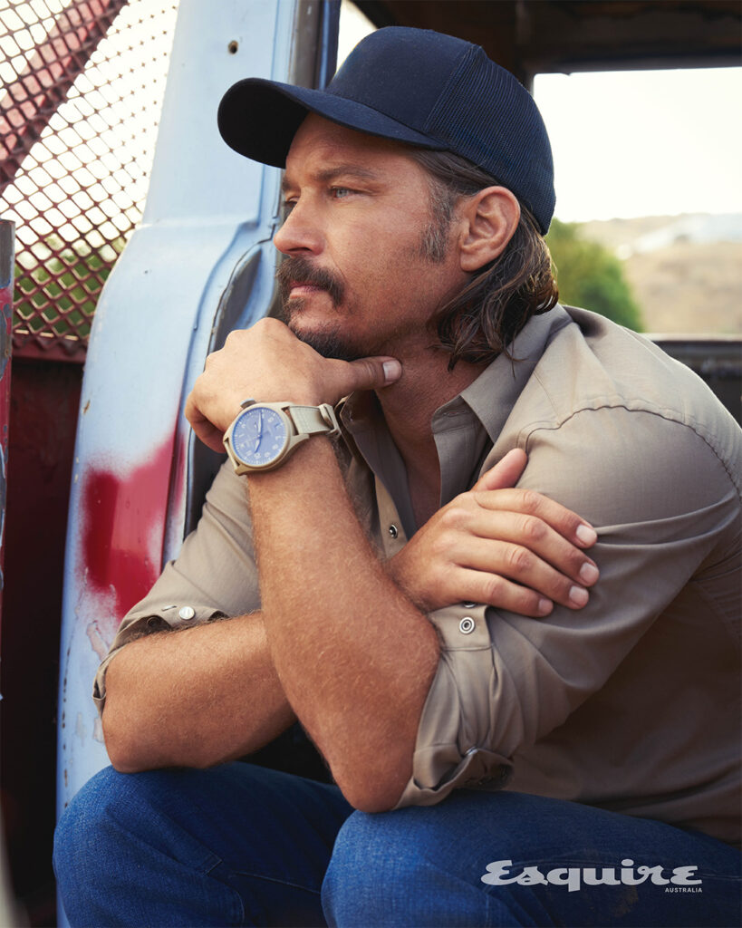 Portrait of adult male wearing a baseball cap and watch, leaning on a distressed blue and red vehicle, in an outdoor setting.