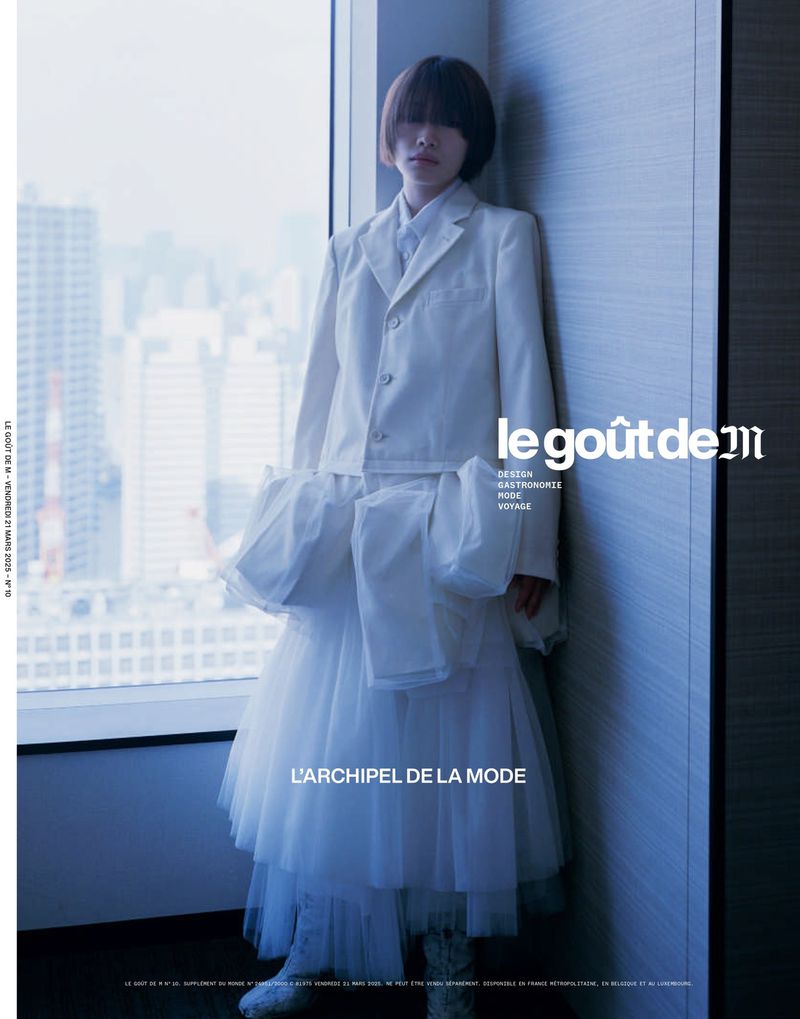 Editorial fashion portrait of a short-haired woman in a white suit with a tulle skirt, posing indoors by a window with a city view.