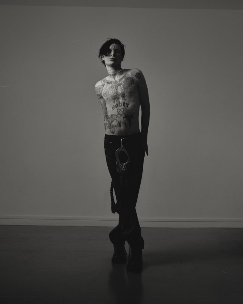 Monochromatic studio portrait of a shirtless, tattooed adult man with dark hair, wearing dark pants and shoes, and accessorized with handcuffs.