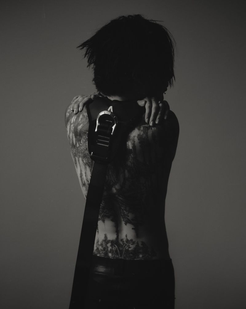 Monochrome back shot of a slender adult with dark hair, tattoos on their arms and back, wearing black pants and a belt.