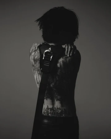 Monochrome back shot of a slender adult with dark hair, tattoos on their arms and back, wearing black pants and a belt.