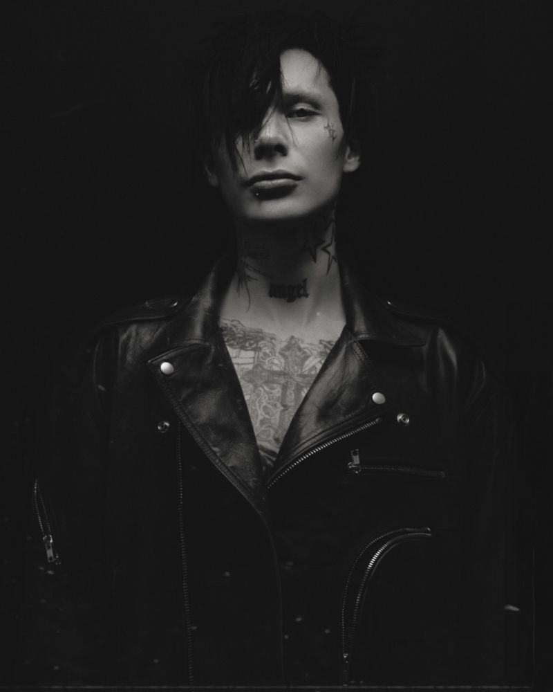 Black and white portrait of a tattooed person with black hair wearing a leather jacket against a dark background. Moody and edgy.