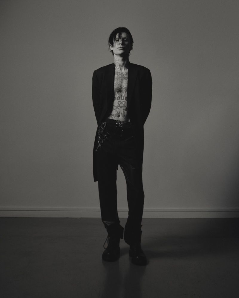 Full-length studio shot of a slender adult male in a black blazer, no shirt, and dark pants, standing in front of a neutral gray background.