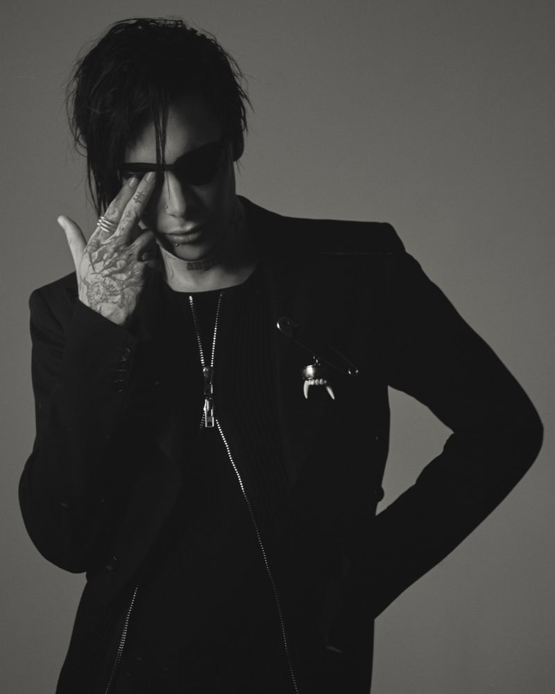 Monochrome portrait of a person with dark hair, sunglasses, and tattoos, wearing a black suit with silver necklace against gray backdrop.