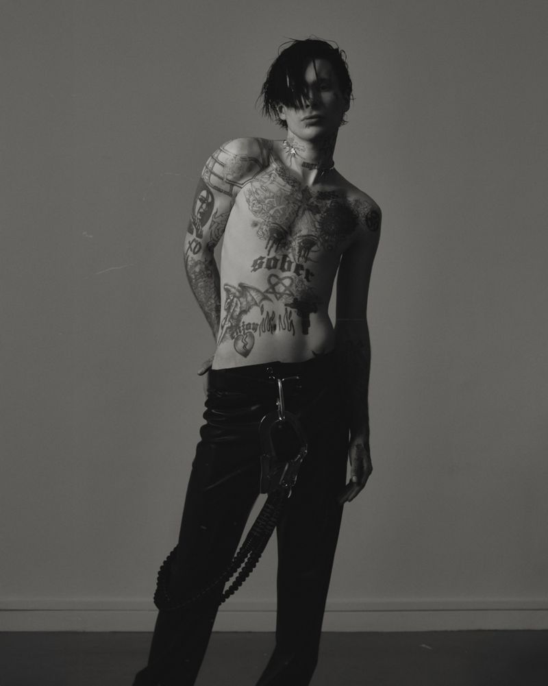 Monochrome studio portrait of a young adult man with dark hair, tattoos, and jewelry, posing shirtless in black pants against a grey backdrop.