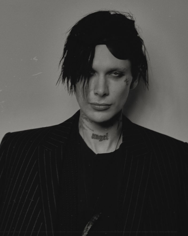 Striking black-and-white portrait of a slender individual with dark hair, tattoos, and a pinstripe jacket against a plain background.