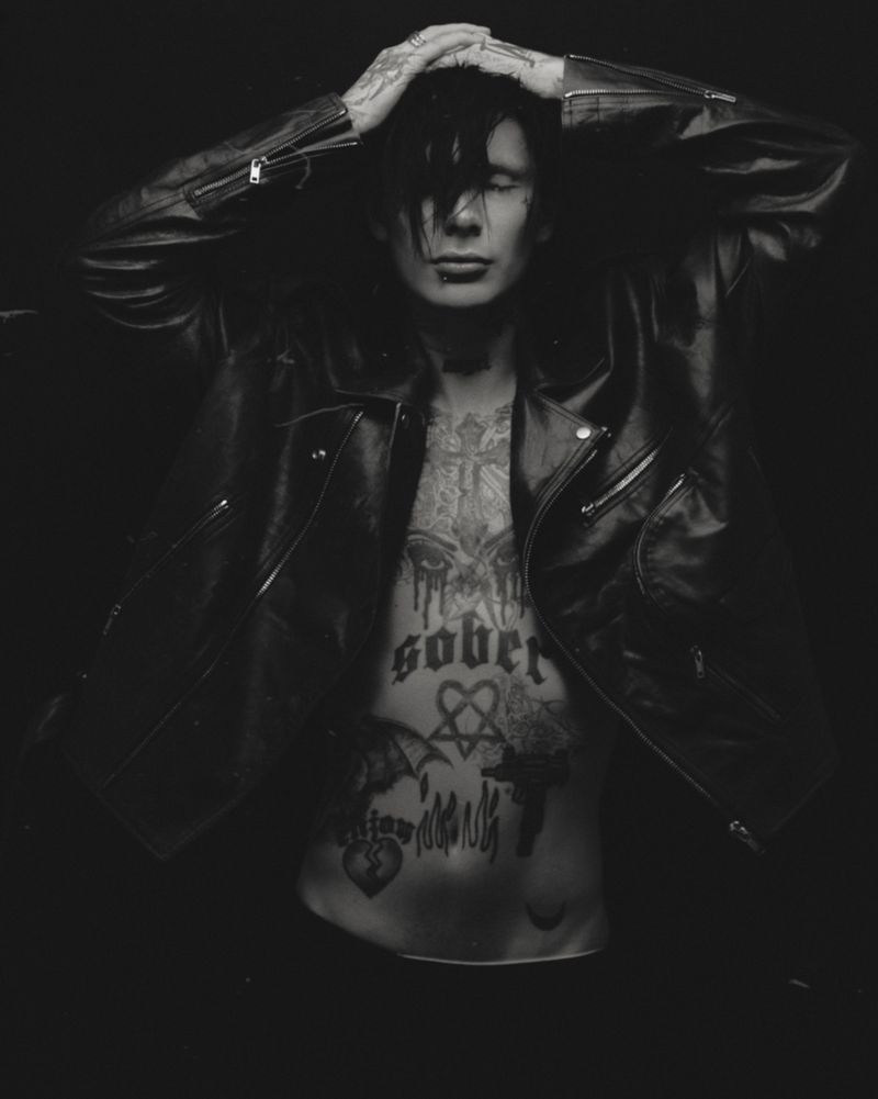 Monochrome portrait of a slender adult with tattoos, wearing a leather jacket and posing with hands above head against a dark background.