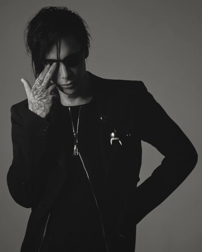 Monochrome portrait of a man in dark sunglasses and a black jacket with hand tattoos raised to his face.