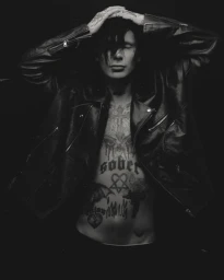 Man with tattooed torso wearing an open black leather motorcycle jacket posing with hands on head in front of a dark backdrop.