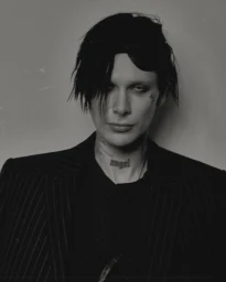 Black and white portrait of 9mice wearing a dark pinstripe blazer with a neck tattoo reading angel against a studio backdrop.