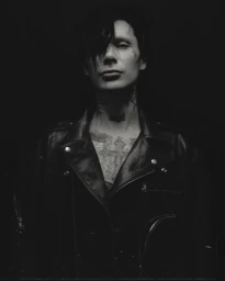 Portrait of a man wearing a black leather jacket with dark hair and chest tattoos, photographed in low key black and white.