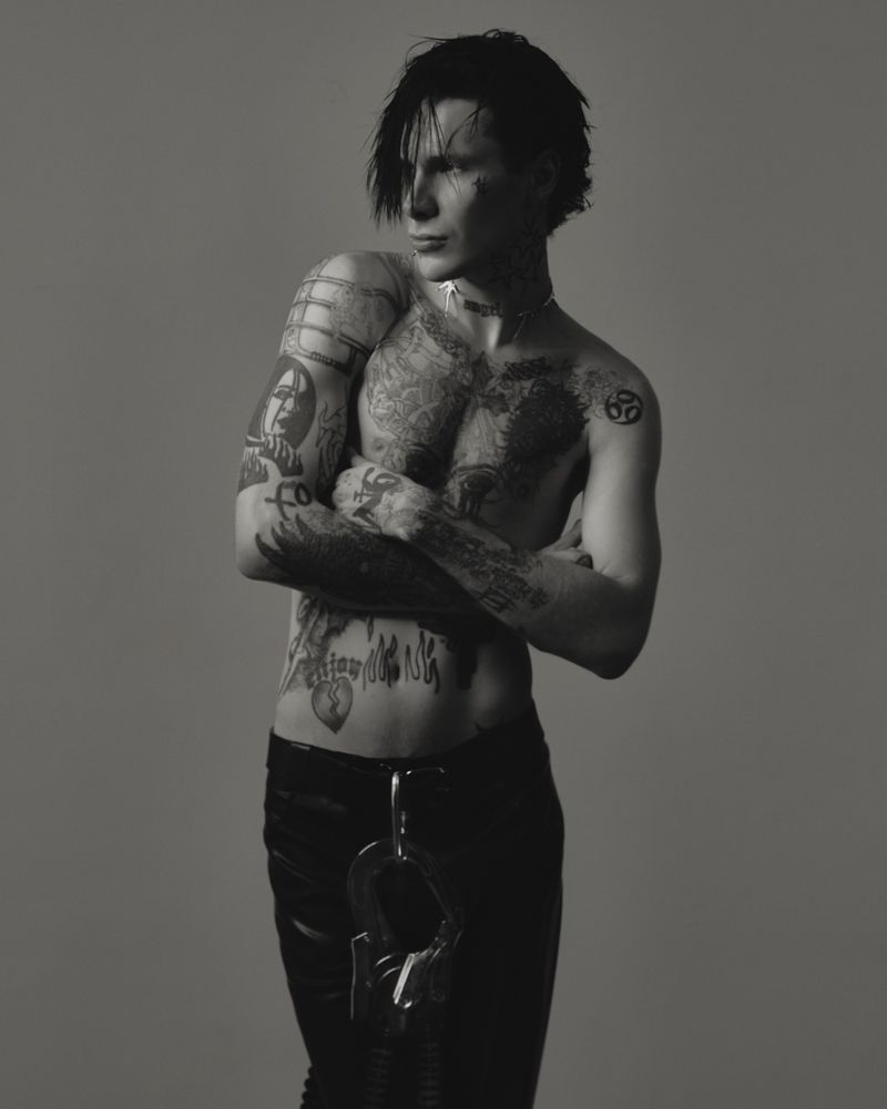 Black and white portrait of tattooed musician 9mice shirtless with crossed arms against a neutral grey studio backdrop.