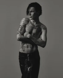 Black and white portrait of tattooed musician 9mice shirtless with crossed arms against a neutral grey studio backdrop.