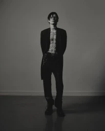 Monochrome portrait of a tattooed man wearing an open blazer and dark trousers against a plain studio background.