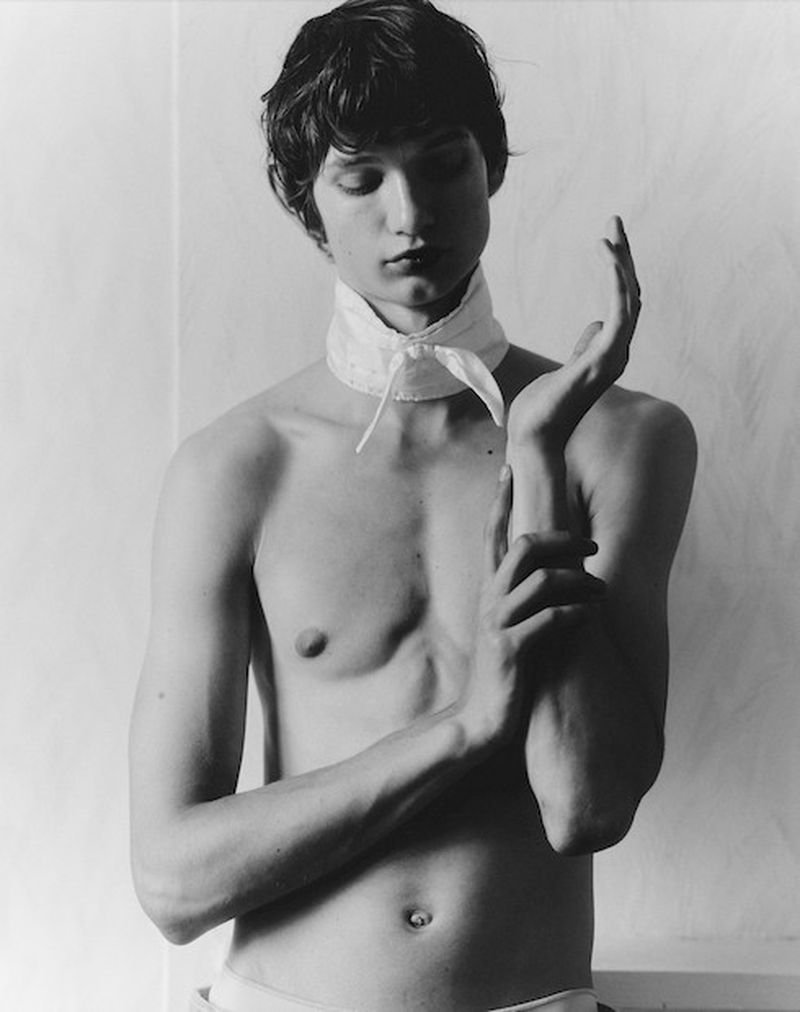 Monochrome portrait of a shirtless young man with dark hair wearing a white scarf, posing indoors against a light wall in a studio setting.