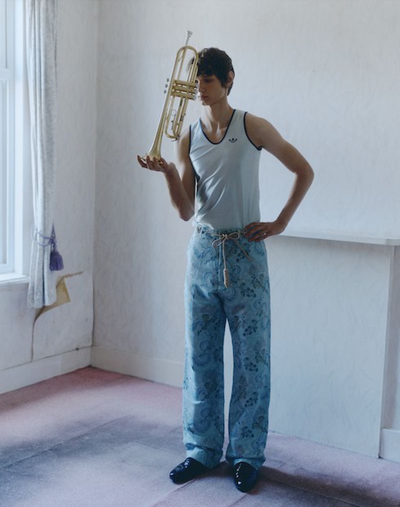 Young adult male model indoors holding a brass trumpet, wearing casual patterned trousers, and posing against a white wall with molding.