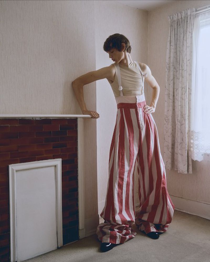 Person poses in a pale beige room wearing a cream tank top with suspenders and red and white striped palazzo pants with black shoes.
