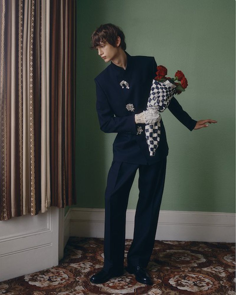 A slender young man in a navy suit holding red roses in a checkerboard wrap, standing in a vintage interior with curtains and carpet.