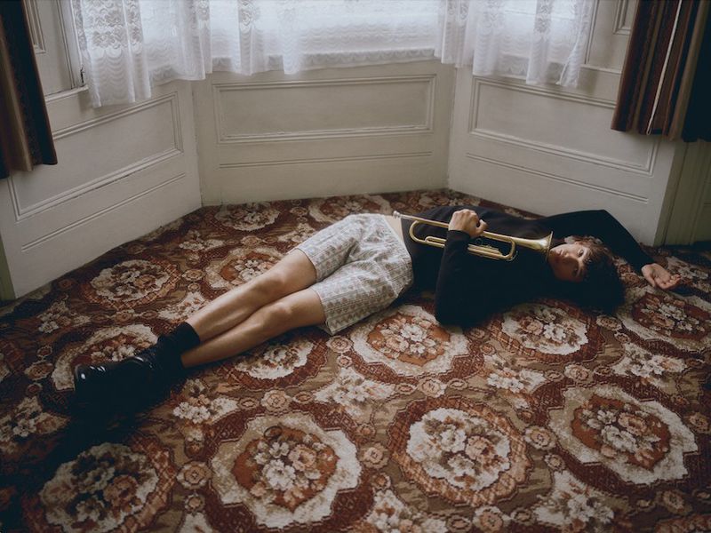 Adult lying on floral rug indoors holding a brass trumpet, wearing a black turtleneck, patterned shorts, socks and shoes, with window light.