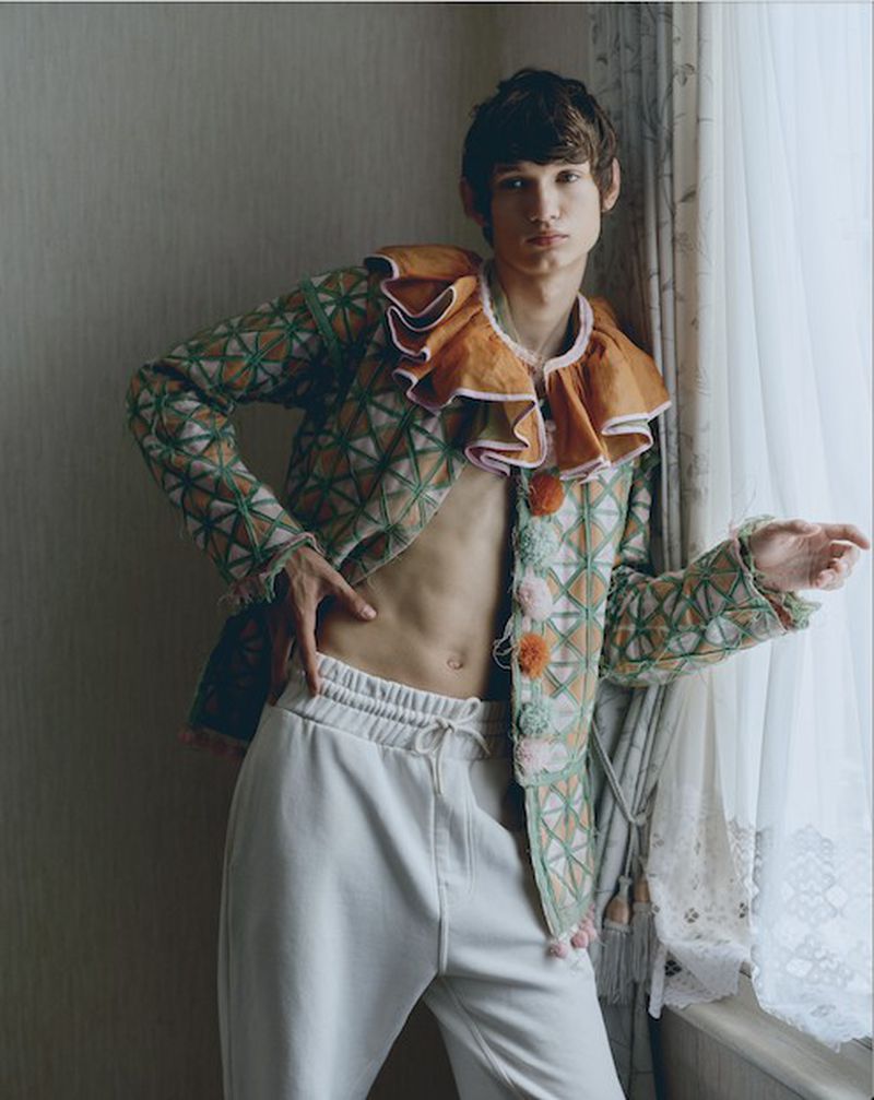 Young male model with patterned jacket poses indoors near a curtain-covered window, showcasing fashion and photographic artistry.