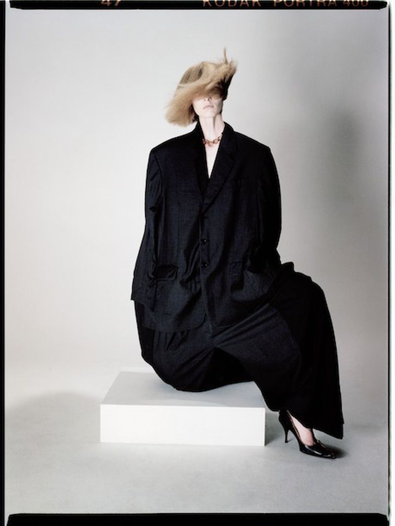 Woman with blond hair sits on a white cube wearing oversized black suit and heels in a studio with white background. Fashion shot.