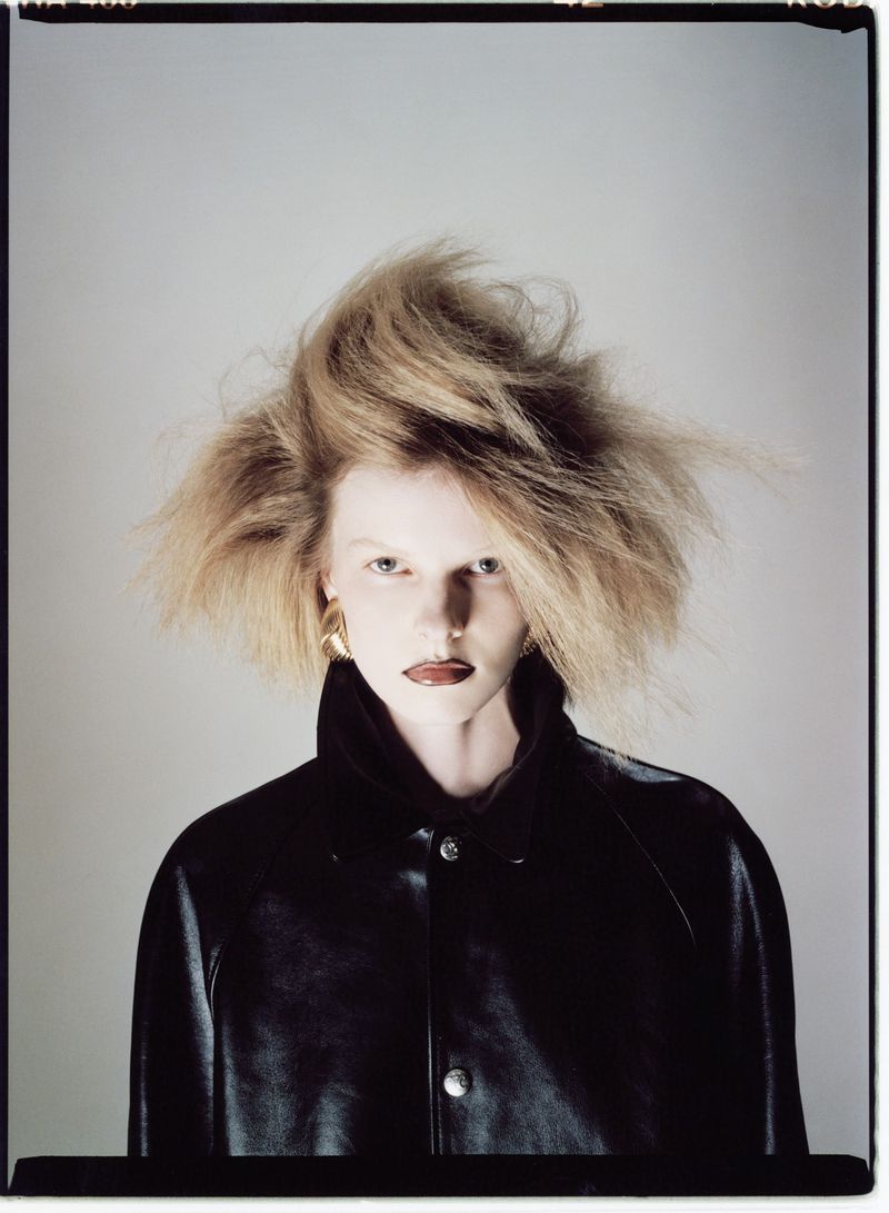 Close-up portrait of a fair-skinned young adult female with blonde teased hair, wearing a black leather jacket against a grey backdrop.