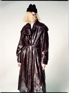 Model Lexy Hearn wearing a glossy dark burgundy patent trench coat and black beanie against a pale studio backdrop.