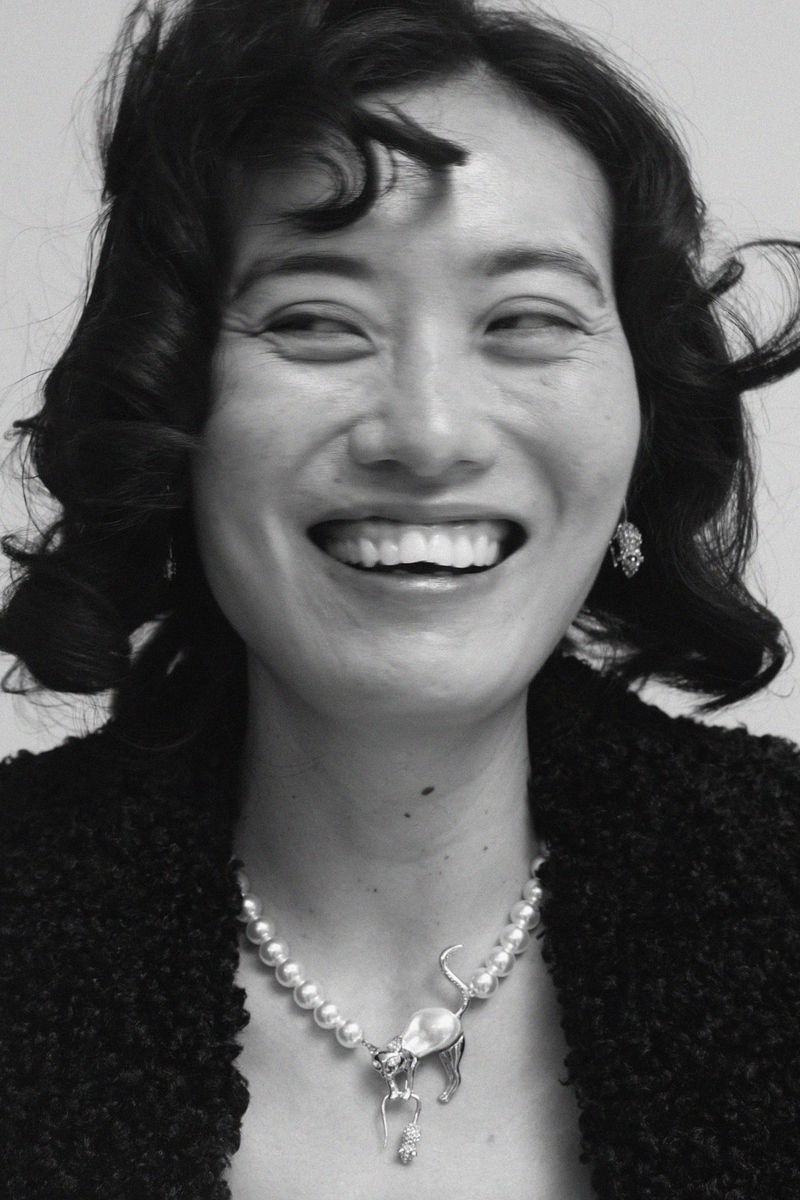 Close-up portrait of a smiling Asian woman with dark curly hair, wearing pearl earrings, a pearl necklace with a cat pendant, and a black sweater.