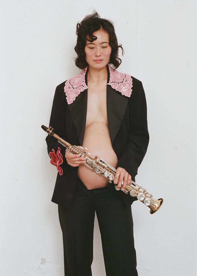 Pregnant woman in a black suit poses against a white wall, holding a saxophone, looking down. Shot in a photo studio.