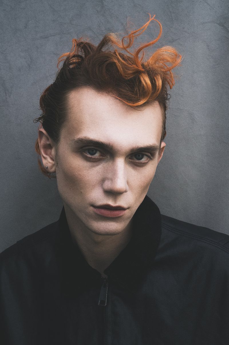 Close-up portrait of a fair-skinned adult male with ginger hair, wearing a black jacket against a grey textured backdrop, looking at the camera.