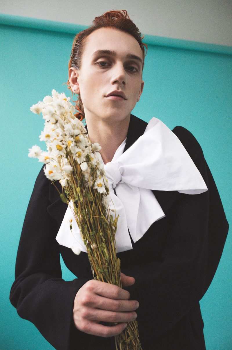 Portrait of a young adult with red hair, wearing a black blazer and a large white bow tie, holding a bouquet of white daisies.
