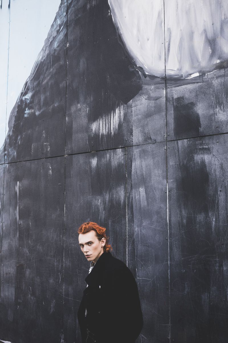An adult male with auburn hair poses in front of an abstract mural on a gray wall, wearing a black jacket, looking at the camera.
