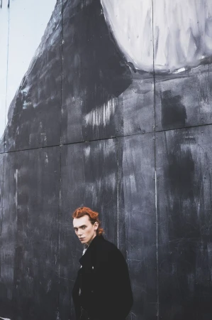 An adult male with auburn hair poses in front of an abstract mural on a gray wall, wearing a black jacket, looking at the camera.