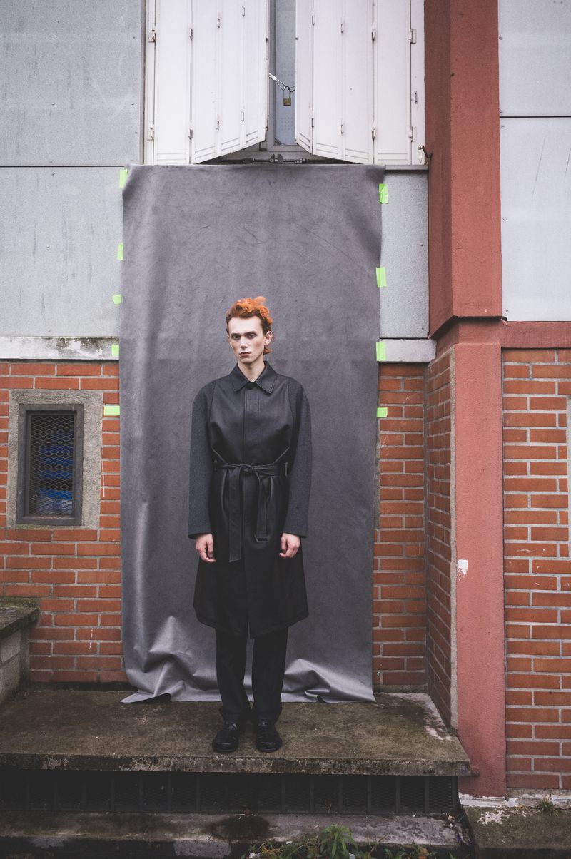 An adult with bright red hair stands in front of a gray backdrop wearing a dark belted coat near a weathered brick building outdoors.