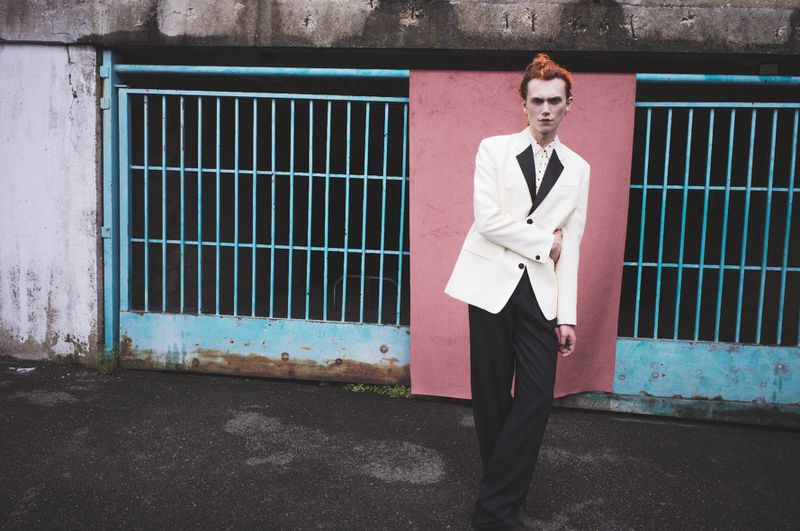Adult male fashion model in white blazer and black trousers stands in urban setting by blue metal gate and pink wall in daylight.