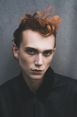Model Ruben Pol with sculpted reddish-orange hair wearing a black jacket against a cool grey textured wall.