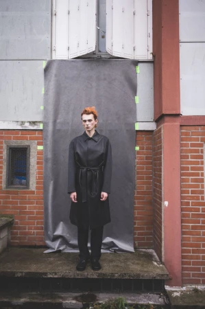Model Ruben Pol with orange hair standing outdoors in a black belted coat in front of a grey taped backdrop.