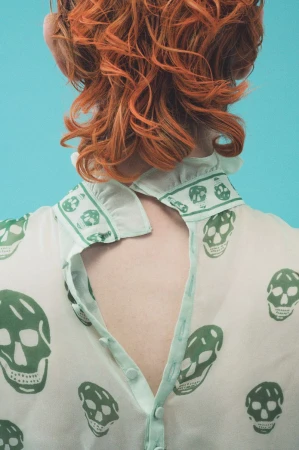 Rear view of a model with bright orange wavy hair wearing a sheer cream blouse patterned with green skulls against a turquoise background.
