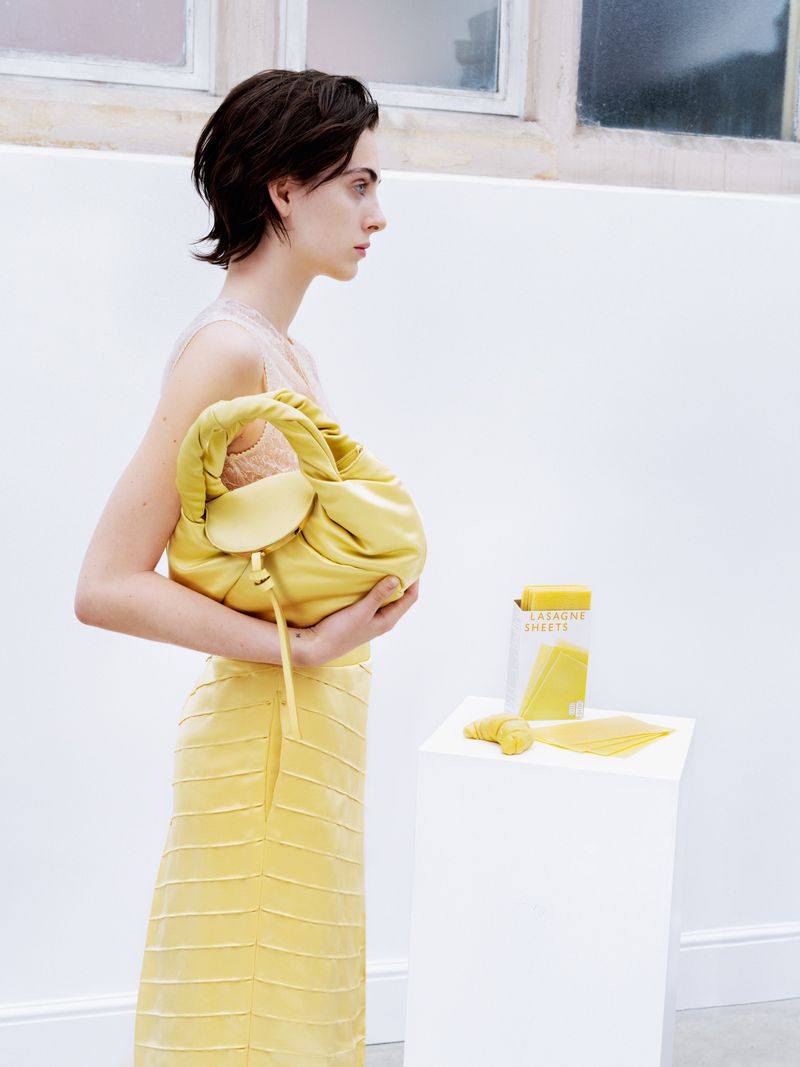Adult woman in profile holds yellow handbag and wears yellow skirt. White background with containers and pasta on white block.
