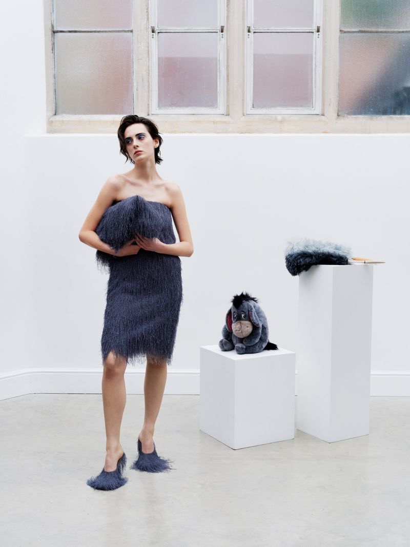 A woman in a fringed dress and heels poses in a bright white space with an Eeyore plush toy, styled for a fashion-forward editorial.