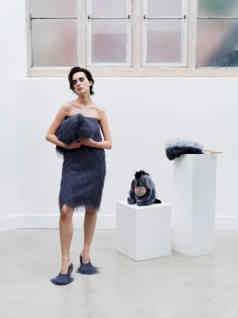 A woman in a fringed dress and heels poses in a bright white space with an Eeyore plush toy, styled for a fashion-forward editorial.