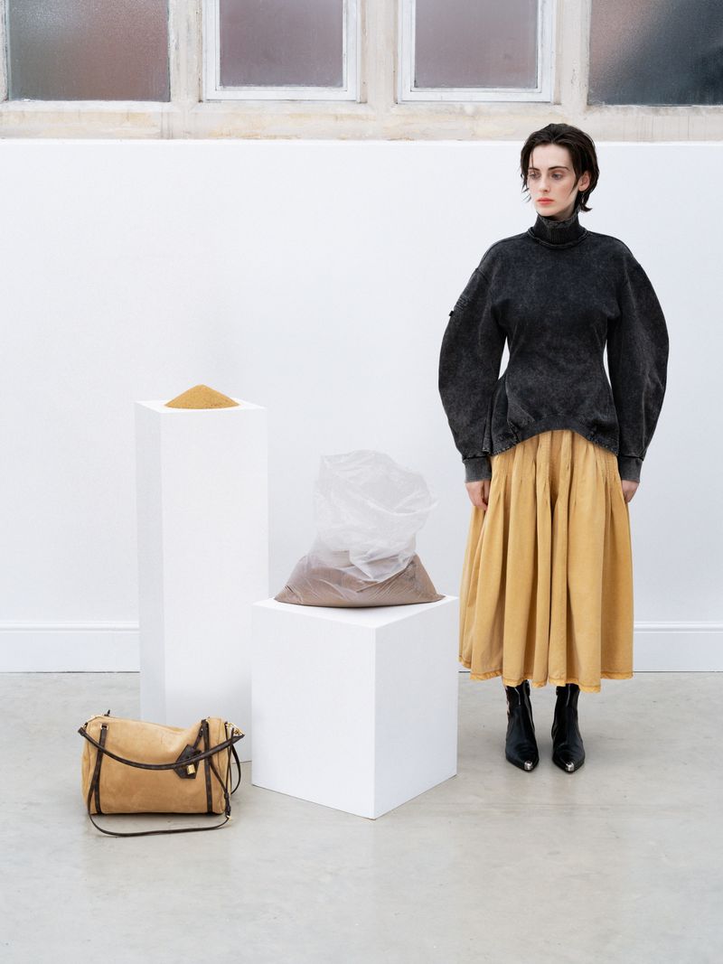 Studio fashion shot of a woman posing in a dark grey top and tan skirt, with a handbag and sand-filled bags on white pedestals.