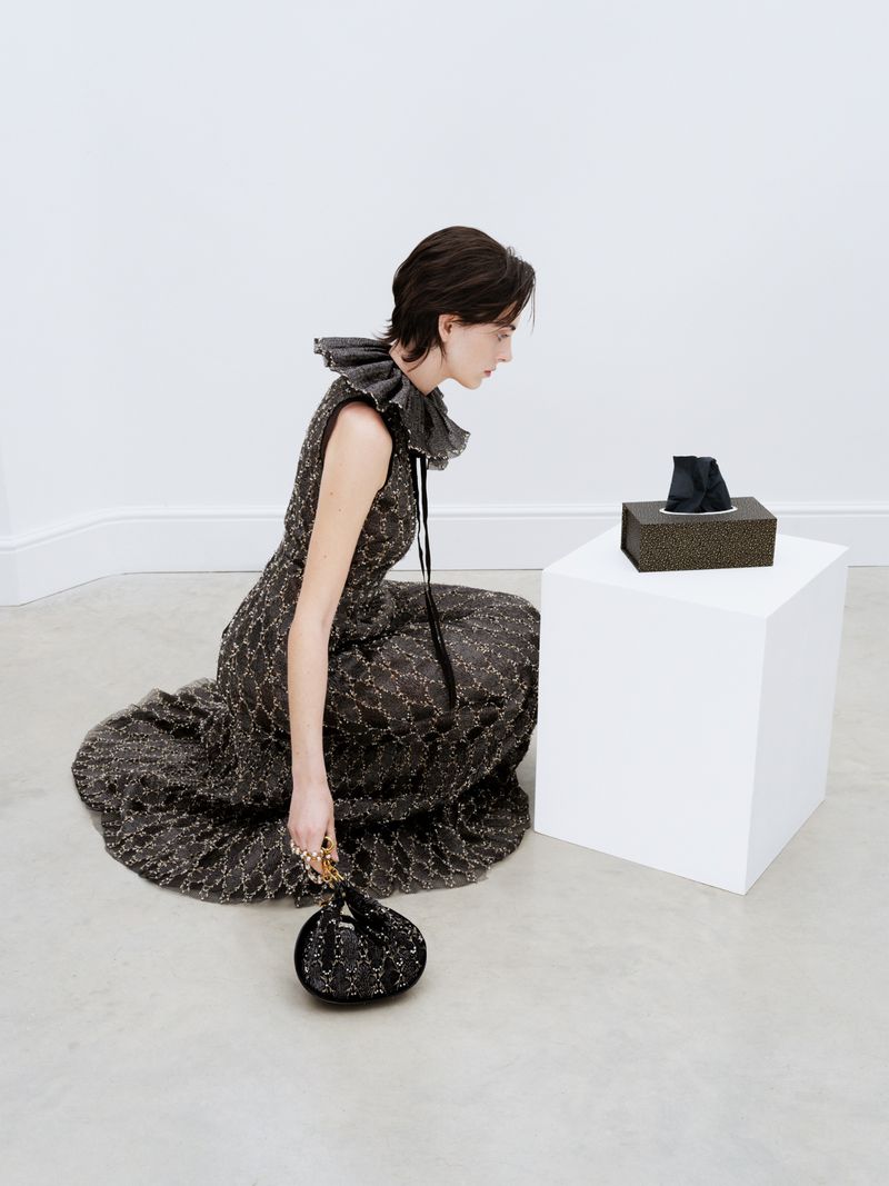 Woman in ruffled collar gown kneeling indoors, looking at a black tissue box on a white cube with a small black handbag at her side.
